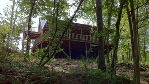 A rustic cabin with a wooden deck and a covered porch, surrounded by dense forest.