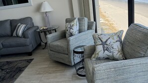 Una sala de estar con un sofá gris, dos sillones, una mesita y una lámpara. Un ventanal grande ofrece vistas a la playa y al océano.