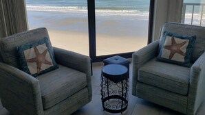 Una habitación con dos sillones grises con cojines estampados con motivos de estrellas, una mesita de noche negra pequeña y vistas a la playa y al océano a través de ventanas amplias.