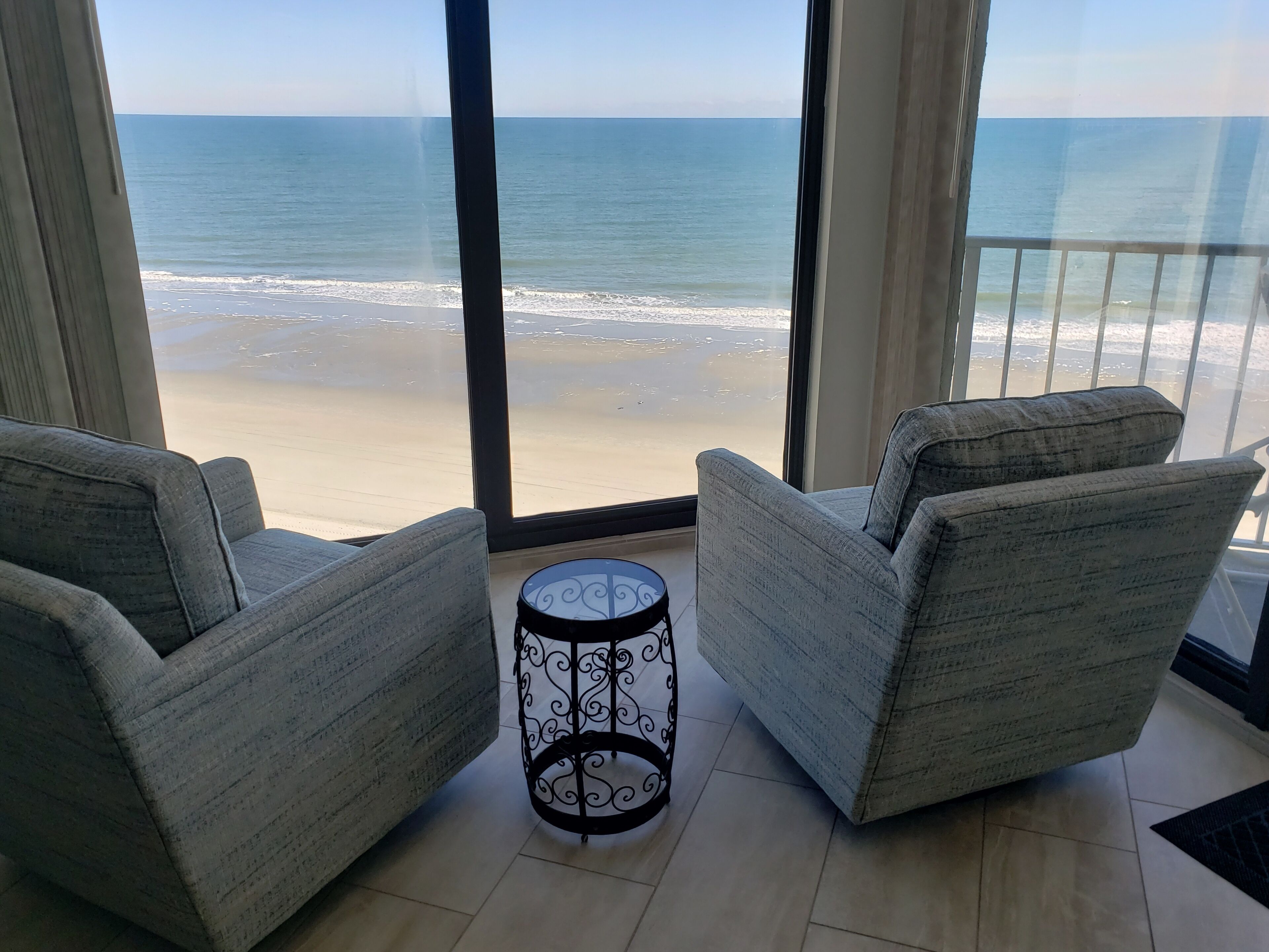 A balcony with two grey armchairs and a small table, overlooking a beach with waves.