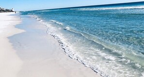 A beach with clear water, a sandy shore, and a few buildings in the background.