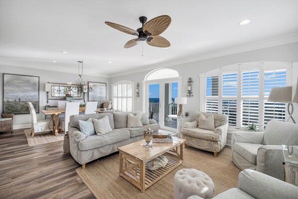 A spacious living room with a large sofa, a coffee table, and a ceiling fan. There is a dining area with a table and chairs, and a view of the ocean through large windows.
