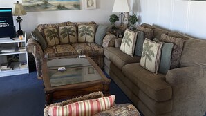 A living room with a sectional sofa, a coffee table, and a patterned ottoman. There are wall-mounted decorations and a lamp on a side table. - HHI Beach & Tennis #AR416, 2BR2BA, top 4th fl, angled Oceanfront condo, 6 max. (Hilton Head Island)