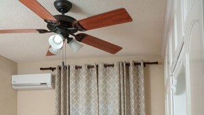 Quarto com cama, ventilador de teto, ar condicionado, luminária e janela com cortinas.