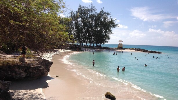 Uma praia com águas turquesa cristalinas, pessoas nadando e caminhando pela orla, e uma torre de salva-vidas ao fundo.