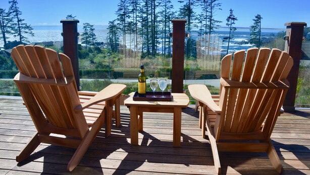 Zwei Holz-Adirondack-Stühle auf einer Holzterrasse, dazwischen ein Tisch mit einer Flasche und zwei Gläsern.