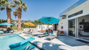 A modern outdoor pool area with lounge chairs and umbrellas, surrounded by palm trees and a house. - Spring into Palm Springs and escape winter! (Palm Springs)
