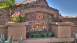 A sign for Canyon Springs with a circular logo, surrounded by plants and a stone wall. - 4 Bedroom Single Story Golf Course Home, Heated Diving Pool + Slide, & Hot Tub  (Phoenix)