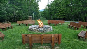 A circular stone fire pit surrounded by wooden benches in a grassy outdoor area with trees. - Westcott House- Privacy, amenities and space all in one beautiful package! (Luray)