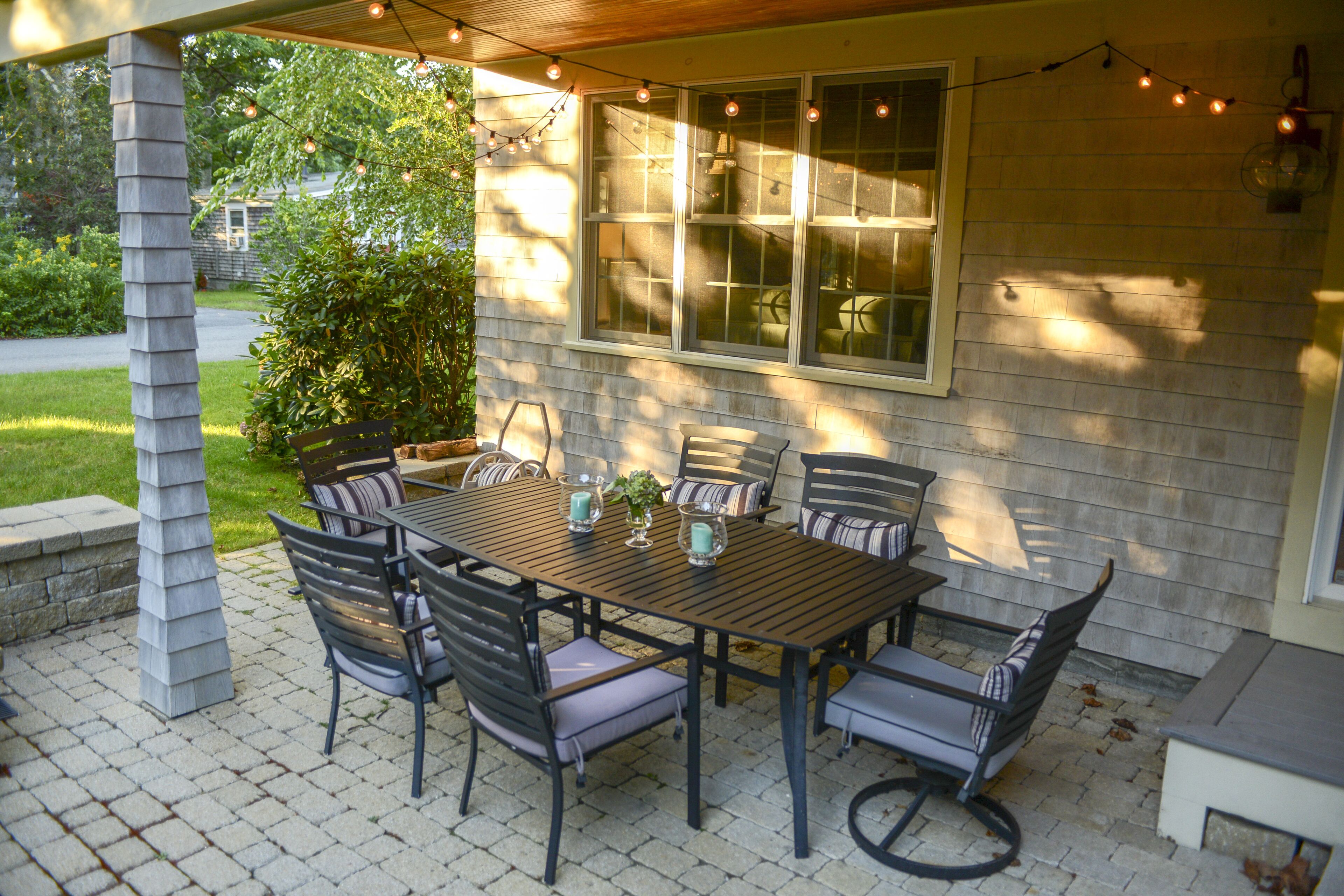 Een overdekt terras met een houten tafel en stoelen, sfeerverlichting en uitzicht op een huis met grote ramen.