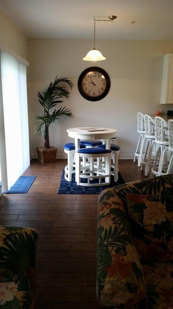 A dining area with a round table and chairs, a potted plant, and a wall clock. - Beautiful Furnished In the Courtyard, Right Beside The Pool On 1st Floor (Gulf Shores)