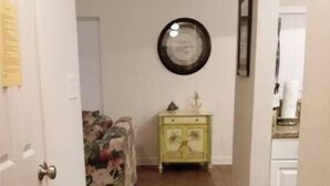 A hallway with wooden flooring, a floral-patterned couch, a yellow cabinet, and a wall-mounted mirror. - Beautiful Furnished In the Courtyard, Right Beside The Pool On 1st Floor (Gulf Shores)