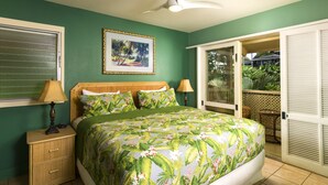 Ein Schlafzimmer mit einem Bett, einem Nachttisch, einer Lampe, einem Deckenventilator, einem Gemälde und einer Schiebetür zu einem Außenbereich mit Pflanzen.