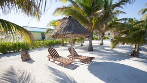 Ein Strand-Loungebereich mit zwei Holzliegen unter einem Reetdach, umgeben von Palmen und einem Sandstrand.