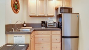 Une cuisine compacte avec des meubles en bois, un évier en acier inoxydable, un four à micro-ondes, un grille-pain et un réfrigérateur. On y trouve des bacs de recyclage et un panneau indiquant les consignes de tri.