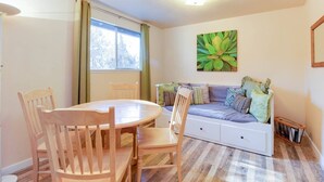 A cozy living room with a round wooden table, two wooden chairs, a sofa with cushions, and a large window with curtains. - The Cascade River Trail Retreat - a short walk to river & downtown! Pets OK! (Bend)