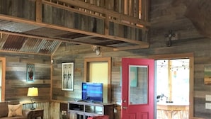 Interior rústico de una cabaña con una escalera de madera, una puerta roja, un televisor y un comedor visible a través de la puerta.