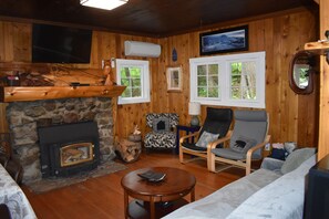 Interior de una cabaña de troncos con chimenea de piedra, paredes de madera, un televisor y un rincón de estar acogedor.