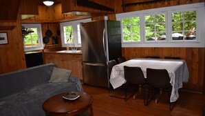 A cabin interior with a sofa, dining table, chairs, a refrigerator, and a kitchen area with a sink and window. - The Cornerstone Cabin - Mount Rainier (Ashford)