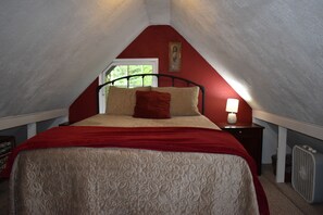 A bedroom with a sloped ceiling, a bed with a red blanket, a nightstand with a lamp, and a radiator. - The Cornerstone Cabin - Mount Rainier (Ashford)