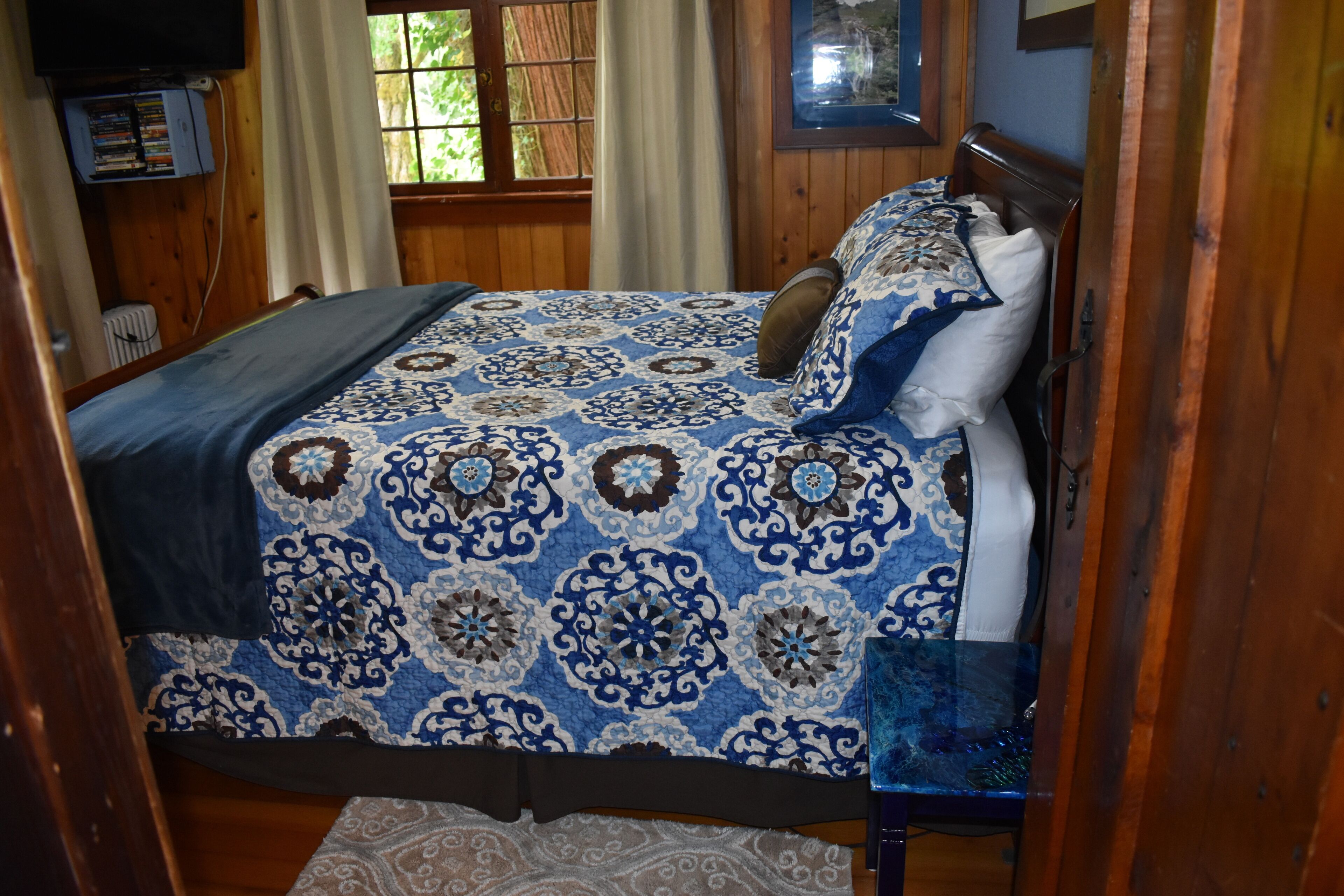 Un dormitorio con cama, paredes de madera, una ventana con cortinas y un televisor en un estante.