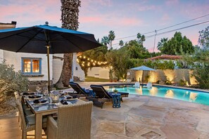 Un área de comedor junto a la piscina, con una mesa preparada para cuatro personas, sillas de descanso y una sombrilla.
