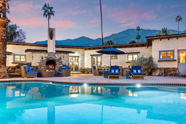 Zona junto a la piscina con chimenea de piedra, mobiliario exterior y vistas a montañas.