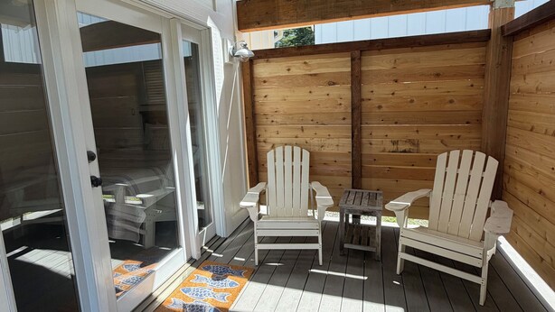 A wooden deck with two Adirondack chairs and a small table, a sliding glass door, and a patterned rug.