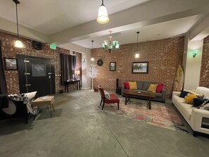 A spacious lobby with a brick wall, a black safe, a red chair, a grey sofa with colorful cushions, and a chandelier.