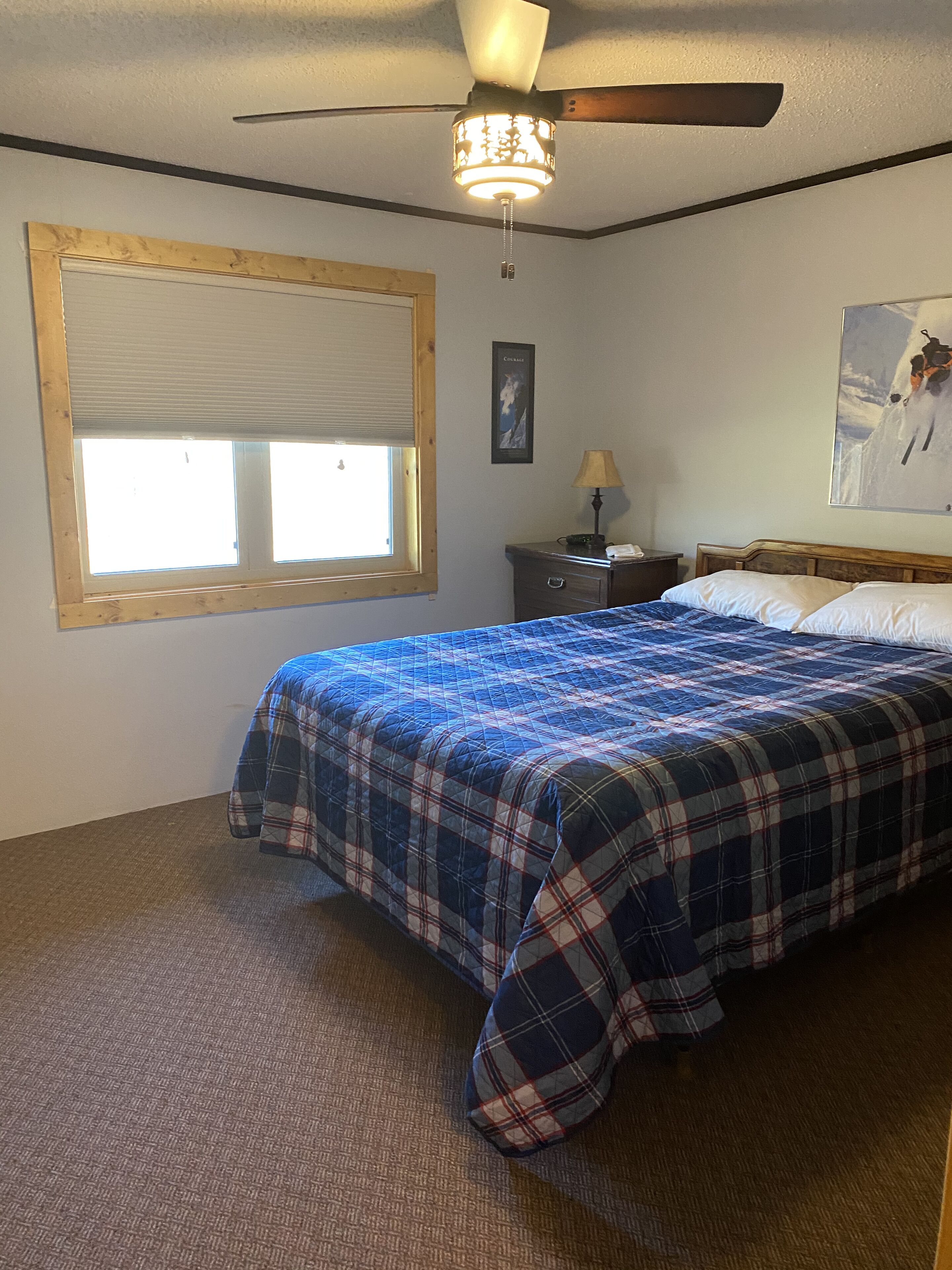 A bedroom with a bed, a ceiling fan, a window with blinds, a nightstand, a lamp, and a painting on the wall.