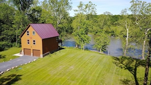 Une cabane en bois à deux étages, avec un toit rouge, un porche à l’avant et un garage, entourée d’une pelouse bien entretenue et d’un lac.