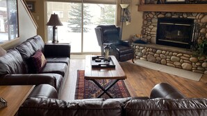 A living room with a stone fireplace, leather sofas, a coffee table, and a large window with a view of a tree. - The Cozy Chalet in Gallatin Range -Warm and Inviting Mountain Chalet (Big Sky)
