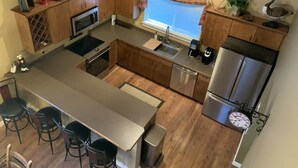 A compact kitchen with wooden cabinets, stainless steel appliances, a central island with bar stools, and a window with blinds. - The Cozy Chalet in Gallatin Range -Warm and Inviting Mountain Chalet (Big Sky)