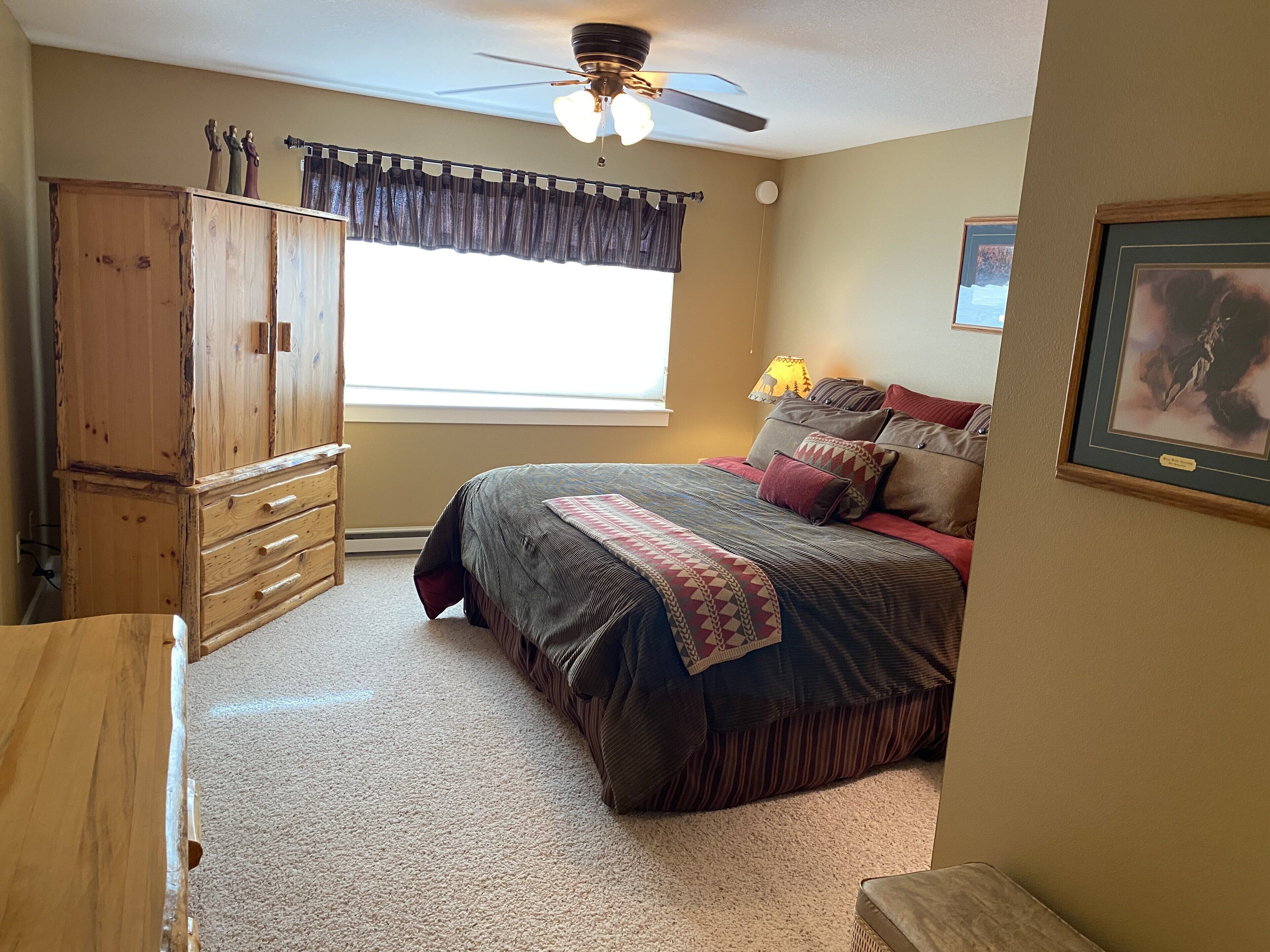Quarto com guarda-roupa de madeira, cama com roupa de cama estampada, ventoinha no teto e janela com cortinas.
