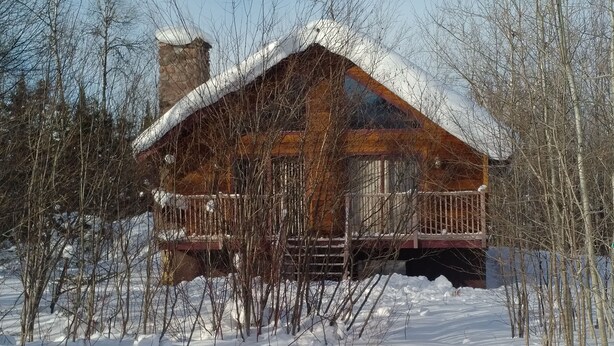 Une cabane en bois, dont le toit est recouvert de neige, entourée d’arbres et de neige.