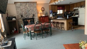 Un salon chaleureux avec une cheminée en pierre, un coin repas avec une table et des chaises, et une cuisine dotée de meubles en bois et d’un distributeur d’eau.