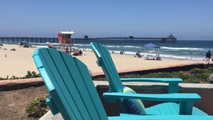A row of turquoise Adirondack chairs with a colorful striped cushion, facing a beach with a pier in the background.
