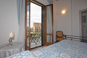 A bedroom with a bed, a chair, a table, and a balcony with a view of buildings.