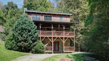 A two-story wooden cabin with a balcony, surrounded by trees and a grassy area.