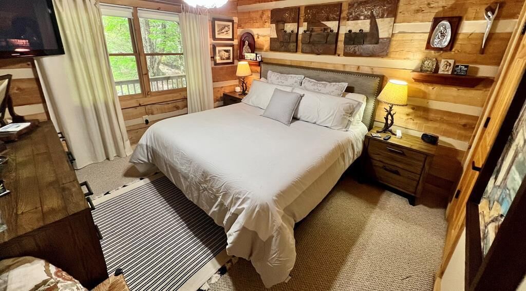 A wooden cabin bedroom with a large bed, two bedside tables with lamps, a TV, and a window with curtains.