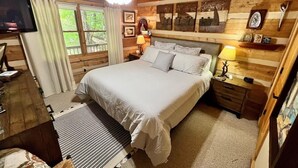 Camera da letto in una baita di legno con un letto grande, due comodini con lampade, una TV e una finestra con tende.