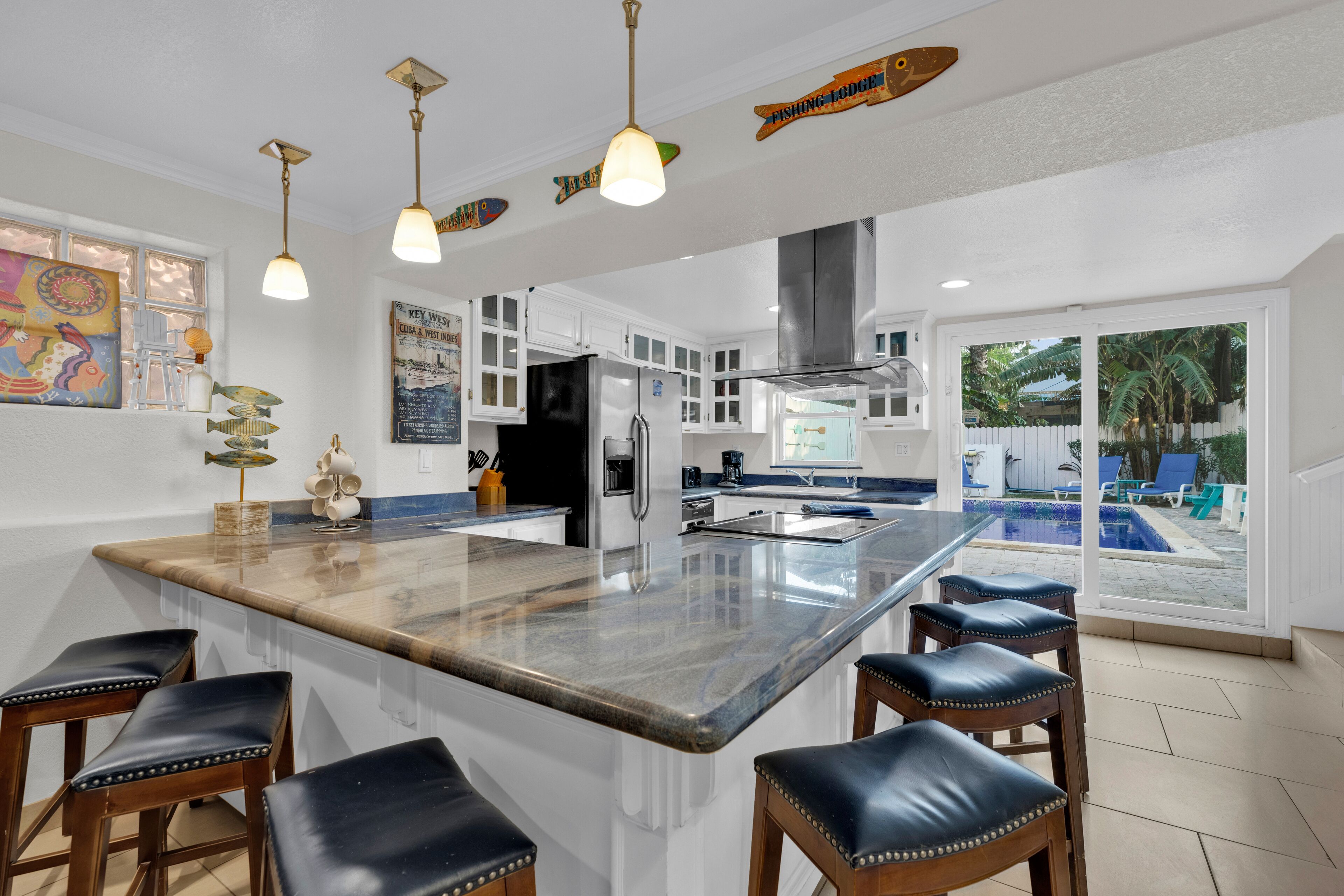 A modern kitchen with a large island, bar stools, pendant lights, and a view of a swimming pool.