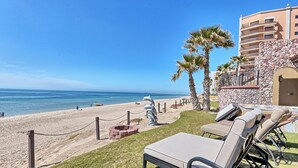 Vista sulla spiaggia con lettini, palme e un edificio in pietra con balconi.