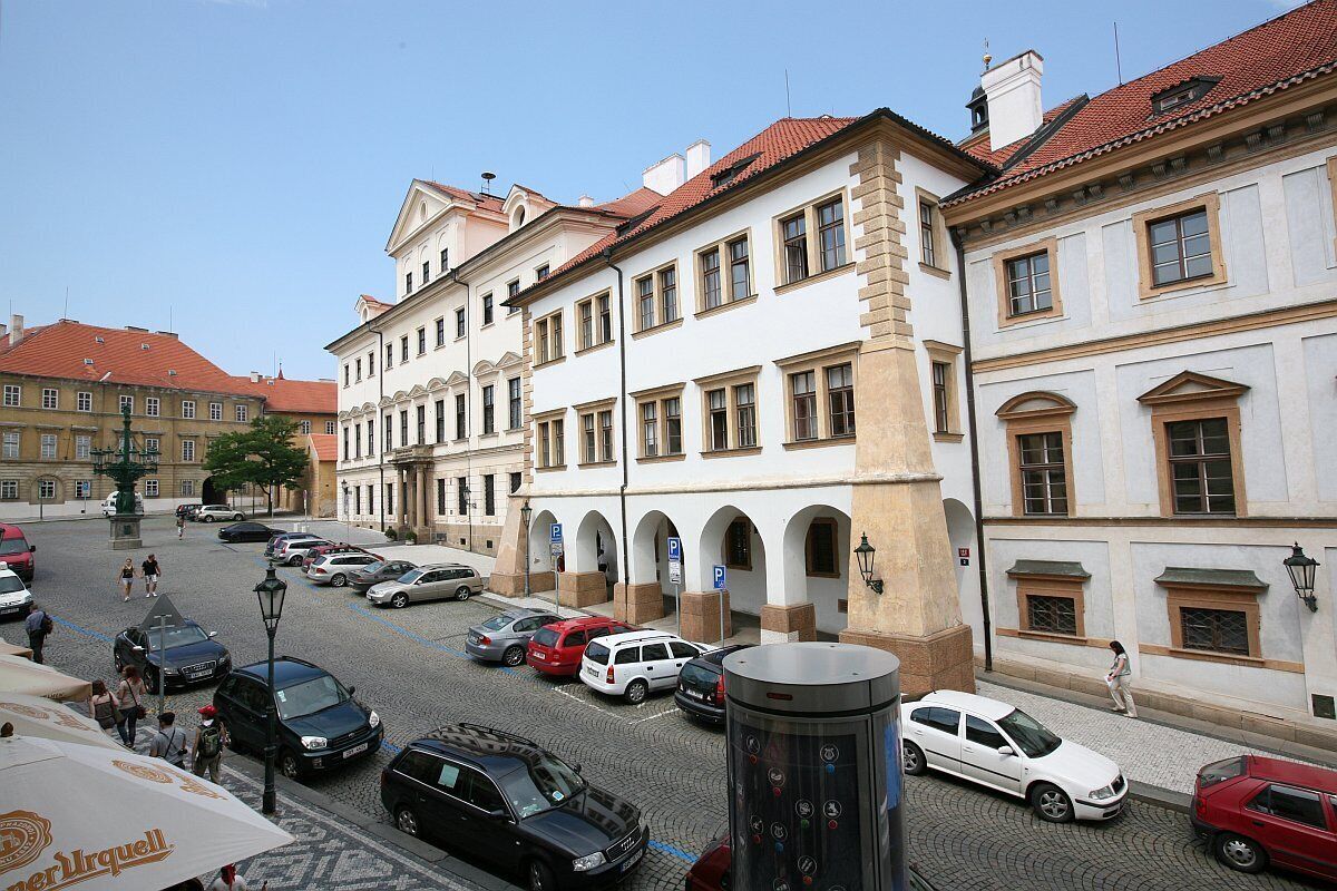Uma rua de paralelepípedos alinhada por edifícios históricos, uma praça pública com uma fonte e carros estacionados.