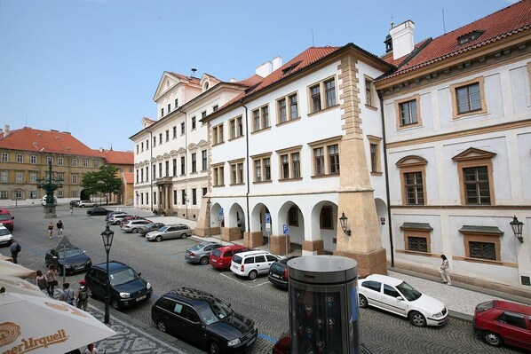 A cobblestone street lined with historic buildings, a public square with a fountain, and parked cars.