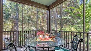 A glass-enclosed balcony with a round table, chairs, and a view of dense foliage.