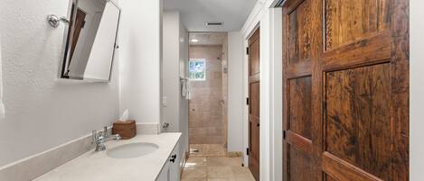 A bathroom with a white sink, a mirror, a wooden door, and a shower area.
