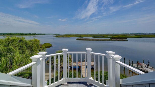 A view from a balcony with a railing, overlooking a calm body of water, greenery, and distant buildings. - Vacation Paradise! Awesome Waterfront Home, Unbelievable View Of Stump Sound! (Surf City)