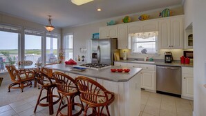 A kitchen with a central island, white cabinets, stainless steel appliances, wicker chairs, and decorative fish on the cabinets. - Vacation Paradise! Awesome Waterfront Home, Unbelievable View Of Stump Sound! (Surf City)