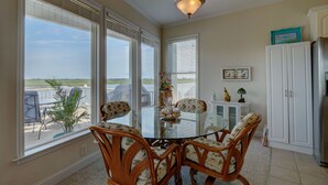 A dining area with a glass table surrounded by wicker chairs, large windows with blinds, a ceiling light fixture, and a cabinet with decorative items. - Vacation Paradise! Awesome Waterfront Home, Unbelievable View Of Stump Sound! (Surf City)